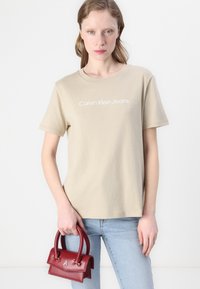 Bež pamukasta majica s kratkim rukavima i bijelim logotipom "Calvin Klein Jeans", u kombinaciji s svijetloplavim trapericama i malom crvenom kožnom torbicom.