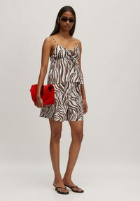 Bruin en witte top met zebra-patronen en spaghettibandjes, gecombineerd met bijpassende shorts. Houdt een rode gestructureerde handtas vast; draagt zwarte zonnebril.