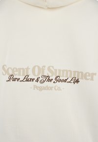 Cream-colored hoodie featuring a textured fabric. The back displays brown and beige graphic text: "Scent Of Summer," "Pure Luxe & The Good Life."