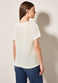 Vit kortärmad stickad topp med en texturerad öppen väv, rund halsringning och en avslappnad passform, tillsammans med blå denimjeans.