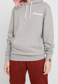 Felpa grigia in tessuto morbido, con logo ricamato bianco, coulisse regolabili e tasca a marsupio. Abbinata a pantaloni rossi.