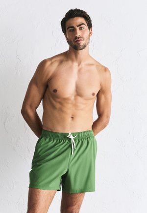Man zonder shirt met donker haar die groene zwemshorts met een witte trekkoord draagt, staat tegen een witte gestructureerde muur.