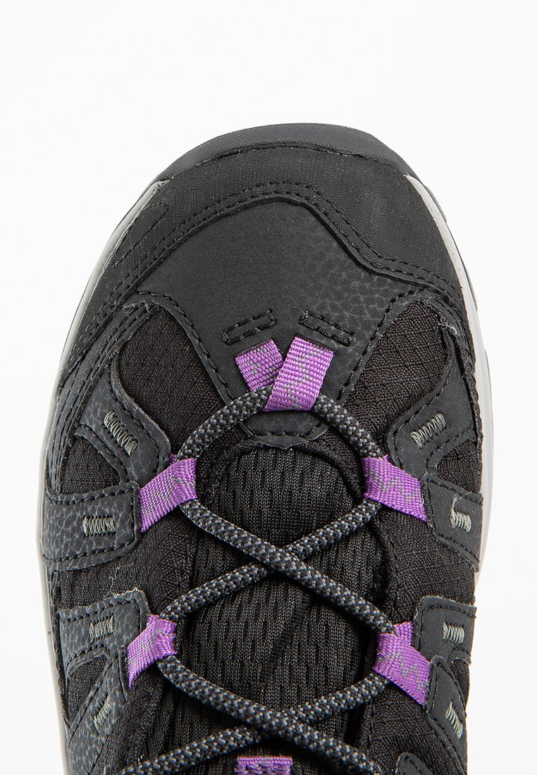 Primer plano de una zapatilla deportiva negra y morada mostrando un tejido texturizado, costuras reforzadas y cordones entrecruzados con lazos morados sobre fondo blanco.