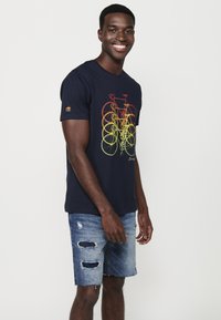 Camiseta de algodón azul marino con un gráfico multicolor de bicicletas superpuestas. El modelo la combina con unos pantalones cortos de mezclilla desgastados.