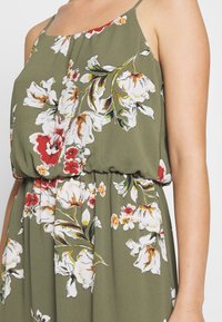 Vihreä kukkamekko, jossa on kootut vyötärön kohdat, spagettiolkaimet sekä kirkkaita valkoisia ja punaisia kukkakuoseja vihreillä yksityiskohdilla.