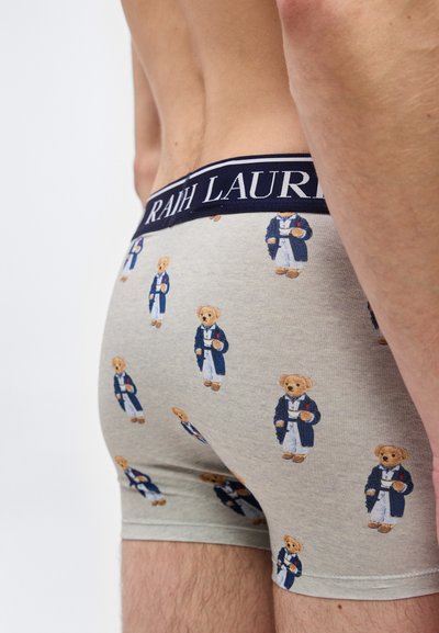 Άνδρας που φοράει ανοιχτόγριμα μποξεράκια με σχέδιο μικρών αρκουδάκια σε μπλε σκούρο παλτό, με μπλε σκούρη μέση που έχει μερικό λευκό κείμενο.