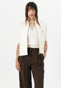 Gilet bianco trapuntato corto con zip frontale e tasca sul petto, abbinato a un top cream a scollo a V e pantaloni marroni a vita alta.