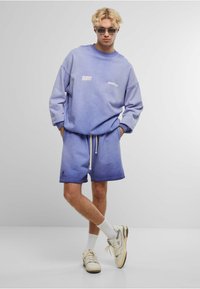 Lichtpaarse sweatshirt en bijpassende shorts, beide met een gradient effect. Het sweatshirt heeft witte tekst, terwijl de shorts een trekkoord en een logo-accent hebben.