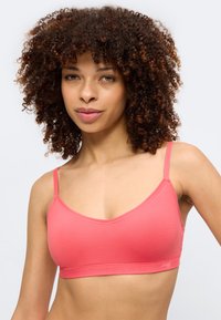 Γυναίκα με σγουρά μαλλιά που φοράει κοραλλί ροζ μπραλέτα, κοιτάζοντας προς τα εμπρός με φόντο ένα απλό φωτεινό περιβάλλον.