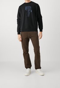 Felpa nera con un design grafico di un personaggio con occhiali da sole, abbinata a pantaloni marroni e sneakers bianche.