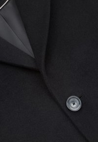 Czarna wełniana tkanina płaszcza o gładkiej fakturze, z klapą z wycięciem i pojedynczym szarym guzikiem. Wnętrze wykończone kontrastową szarą podszewką.