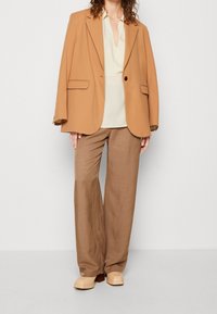 Blazer beige avec des revers crantés, blouse beige et pantalon loose marron. Le modèle porte des chaussures à plateforme beiges épaisses. Tissu lisse ; coupe décontractée.