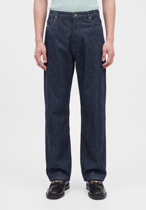 Homme portant un jean droit bleu foncé, des mocassins noirs avec des détails de chaîne dorée, et un haut sans manches côtelé bleu clair.