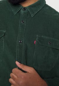 Chemise en velours côtelé vert foncé avec un col à revers, des boutons noirs et deux poches avant. Présente une texture distinctive à côtes verticales.