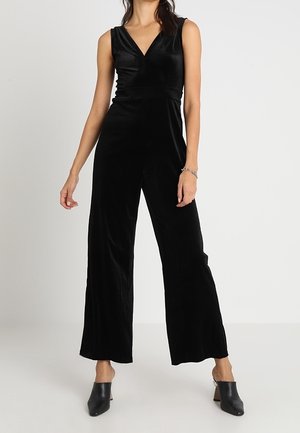 Femme portant une combinaison sans manches en velours noir à jambes larges et des mules noires à talons, sur un fond gris uni.