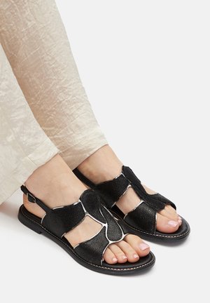 Sandali neri con tomaia strutturata caratterizzata da incisioni e bordi bianchi, su suola piatta, abbinati a pantaloni chiari.