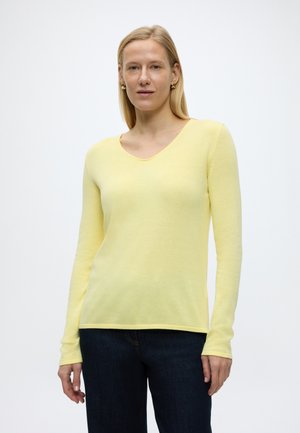 Žena s rovnými blond vlasmi, ktorá má zlaté kruhové náušnice, svetložltý sveter s dlhým rukávom a výstrihom do V a tmavomodré džínsy, na bielom pozadí.