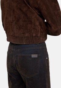 Brązowa zamszowa kurtka cropped z teksturowanym wykończeniem, zestawiona z ciemnymi dżinsami z metką na tylnej kieszeni i dopasowanym krojem.
