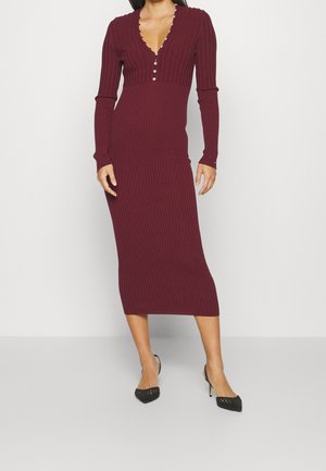 Vrouw draagt een lange mouw, geribbelde, bordeauxrode midi-jurk met een V-hals en knoopdetails, gecombineerd met zwarte pumps met spits toelopende neus.