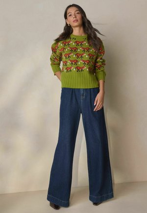 Grüner Strickpullover mit Fuchs-Muster in Rot und geripptem Saum, kombiniert mit blauen weit geschnittenen Jeans und dunklen Absatzschuhen.