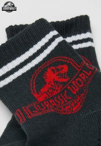 Calze nere con il logo rosso di Jurassic World, caratterizzate da strisce bianche sulla parte superiore. Texture a maglia con un logo che rappresenta un dinosauro.