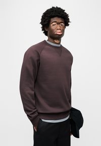 Σκούρο καφέ φούτερ με στρογγυλή λαιμόκοψη και μανίκια ράγκλαν, σε συνδυασμό με ανοιχτό γκρι εσώρουχο και μαύρο μπέιζμπολ καπέλο στο χέρι.