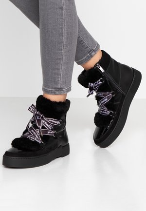 Mustat tekoturkiksella vuoratut nilkkakengät, joissa on musta patenttinen nahkaosa, koristeelliset purppuranväriset nauhat tekstillä ja paksu musta pohja.