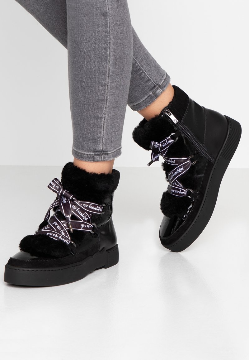 Mustat tekoturkiksella vuoratut nilkkakengät, joissa on musta patenttinen nahkaosa, koristeelliset purppuranväriset nauhat tekstillä ja paksu musta pohja.