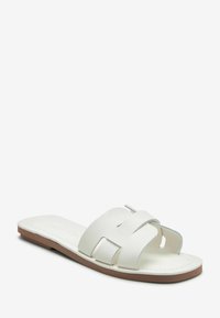 Sandal de piel blanca con diseño de trenzado, suela plana y suela de goma marrón claro. Textura suave con un mínimo de detalles.