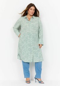 Un vestito camicia verde chiaro con un motivo astratto bianco testurizzato; maniche lunghe; due tasche laterali; abbinato a jeans blu e sandali con tacco.