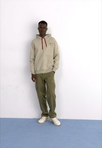 Sudadera con capucha beige con cordones burdeos y bolsillo frontal, combinada con pantalones cargo oliva y zapatillas blancas, frente a un fondo sencillo.