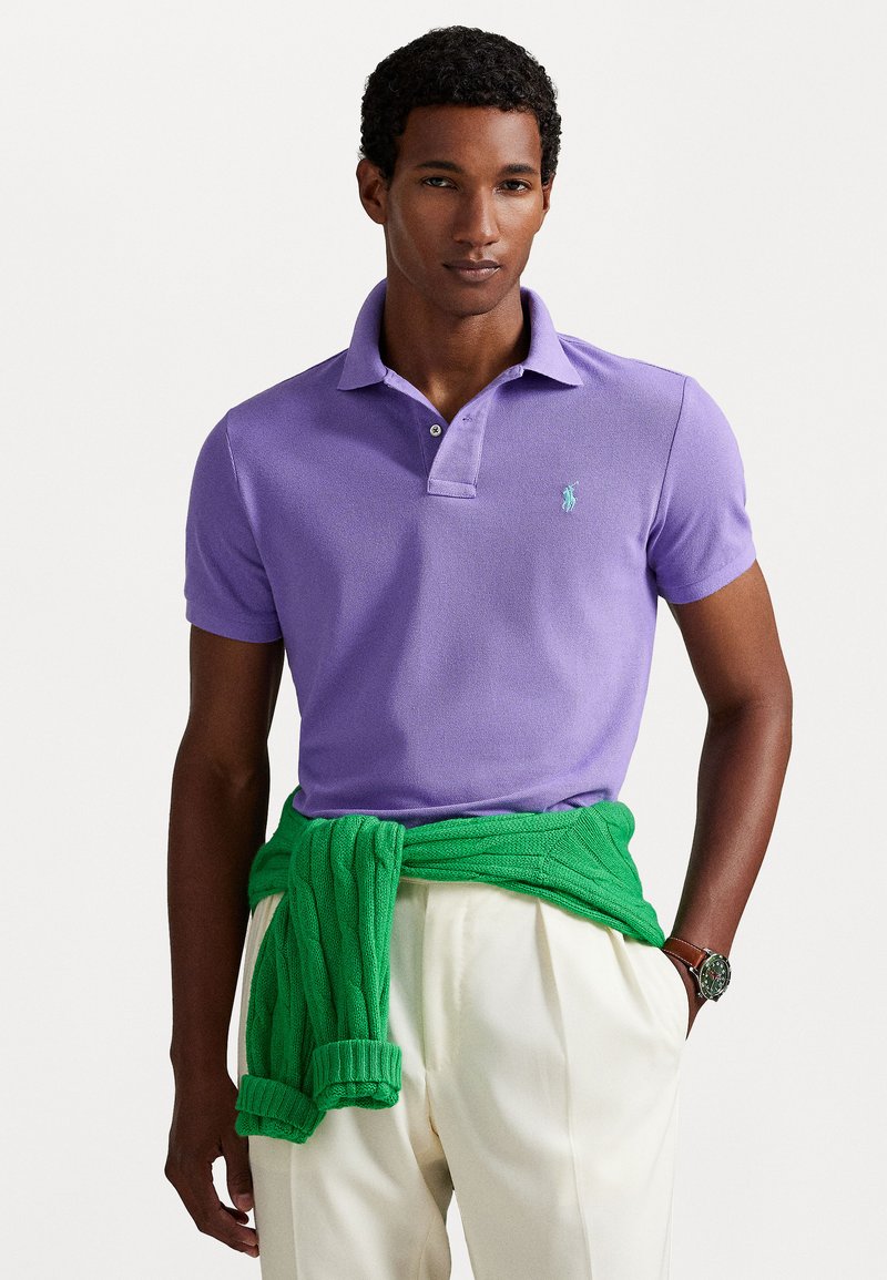 Vijolična polo majica z teksturirano tkanino, opremljena z majhnim akvamarinskim logotipom. Zelena pletena jopica z vrvico na pasu, v kombinaciji z svetlimi hlačami.