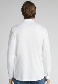 Man met bruin haar, gekleed in een witte lange mouwen shirt en blauwe jeans, met zijn rug naar de camera toe tegen een effen lichtgrijze achtergrond.