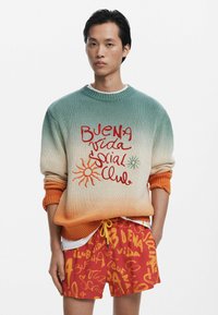 Sweter dzianinowy w gradientowym kolorze od teal do kremu, z pomarańczowymi mankietami, ozdobiony czerwonym i zielonym haftem oraz motywami słońca. W połączeniu z czerwonymi szortami w wzory.