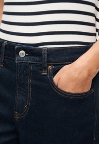 Άτομο που φορά σκούρο μπλε τζιν με το χέρι στην μπροστινή τσέπη και λευκό πουκάμισο με μαύρες οριζόντιες ρίγες.