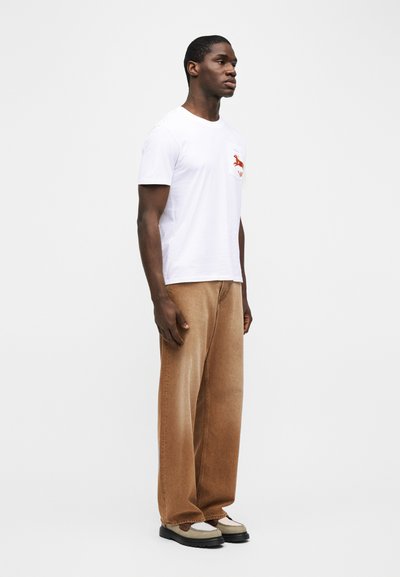 Άντρας που στέκεται προφίλ, φορώντας λευκό μπλουζάκι με κόκκινο λογότυπο ελαφιού, χαλαρό καφέ παντελόνι και μπεζ και μαύρα παπούτσια.