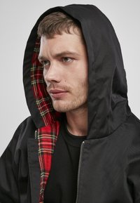 Zwarte hooded jack met een rood en groen geruite voering. Voorzien van een ritssluiting en een relaxte pasvorm. Gekocht boven een zwart shirt.