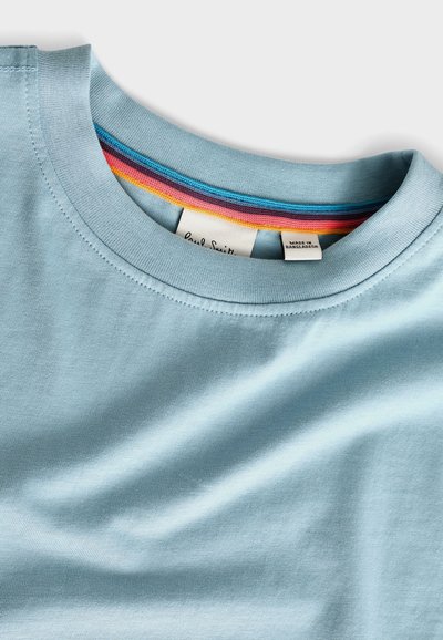 Gaiši zils kokvilnas T-krekls ar apaļu kakla izgriezumu, kura apakšmalā ir strīpota apakšmala rozā, zila un dzeltena krāsā. Uz etiķetes ir redzama zīmola informācija.