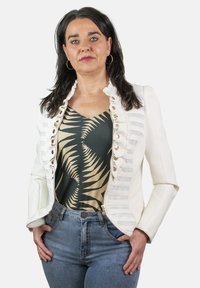 Chaqueta de cuero sintético blanca con detalles de volantes, combinada con una blusa de estampado verde y beige y jeans de mezclilla azul. Los accesorios incluyen pendientes de aro.