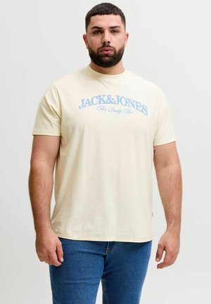 Man met baard die een crèmekleurig "JACK & JONES For Daily Use" t-shirt en blauwe spijkerbroek draagt, staand tegen een effen witte achtergrond.