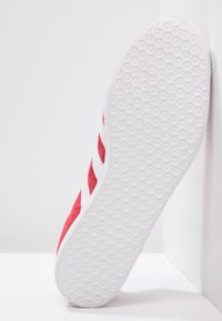 Červené semišové tenisky s bílou gumovou podrážkou, vyznačující se třemi bílými pruhy po straně a texturovaným hexagonálním vzorem na podrážce.