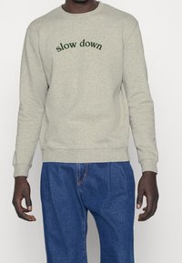 Felpa grigia con testo "slow down" in verde scuro. Collo rotondo, polsini e orlo a costine. Indossata con pantaloni in denim blu.