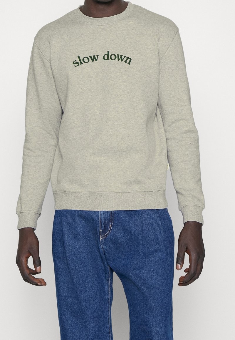 Felpa grigia con testo "slow down" in verde scuro. Collo rotondo, polsini e orlo a costine. Indossata con pantaloni in denim blu.