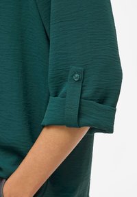 Teal skjortärm med en texturerad tyg, som har en rullad manschett och en matchande knappdetalj. Ren, avslappnad design med en mjuk finish.