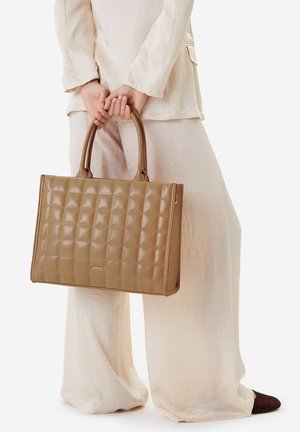 Persona con atuendo beige claro sosteniendo un gran bolso tote de cuero marrón acolchado con doble asa y la marca "JENNY".