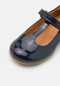 Lakkinen tummansininen Mary Jane -kenkä, jossa on vaaleanruskea sisus ja beige pohja, varustettuna T-nauhoilla ja ompeluyksityiskohdilla valkoisella taustalla.