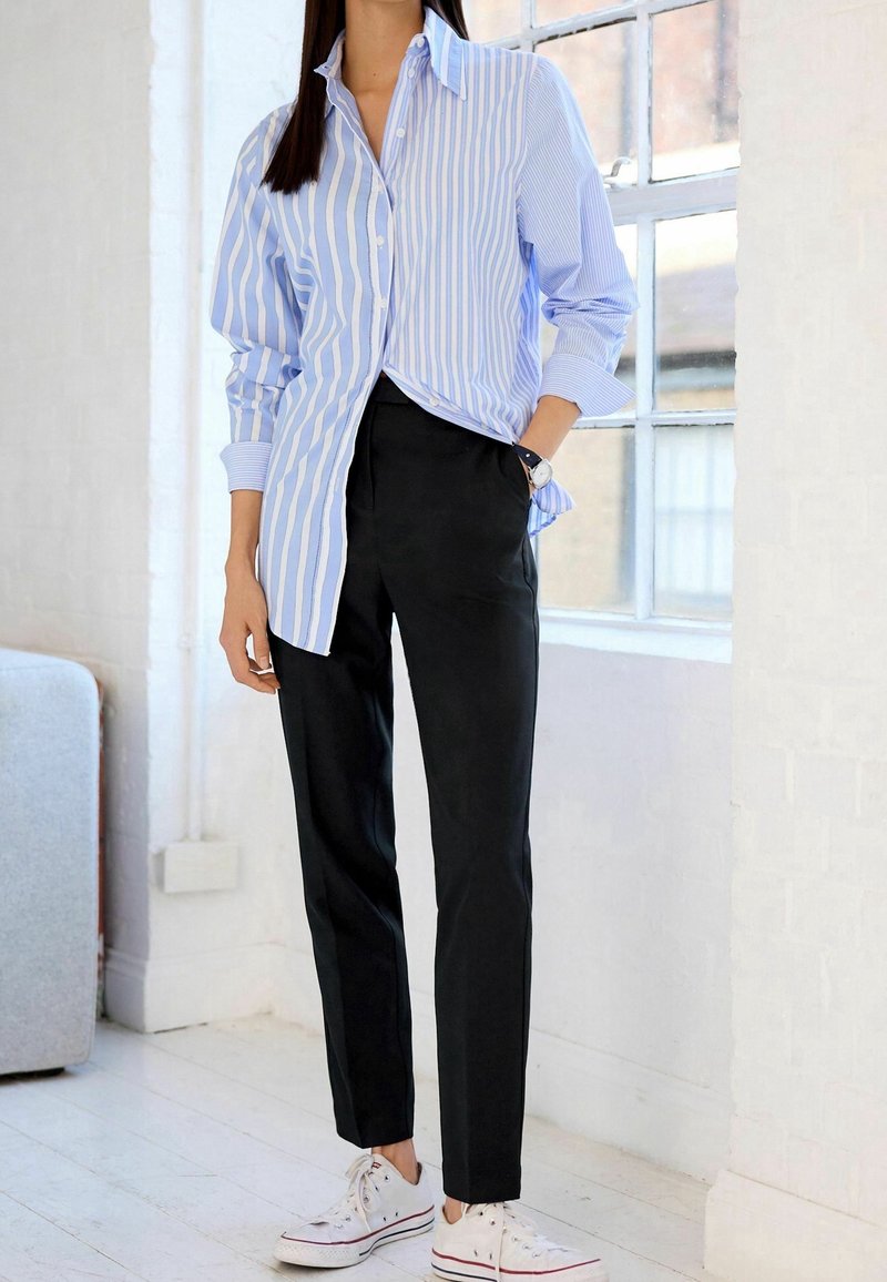 Femme portant une chemise à rayures bleues et blanches boutonnée, pantalon noir, baskets blanches, debout dans une pièce lumineuse près d'une fenêtre.