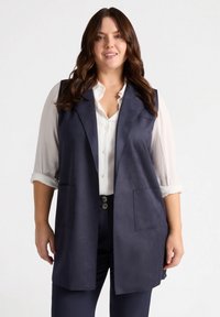 Marineblau ärmellose Weste aus Faux Wildleder mit Reverskragen, zwei Fronttaschen und offenem Vorderteil. Getragen über einem weißen Hemd mit Knöpfen.