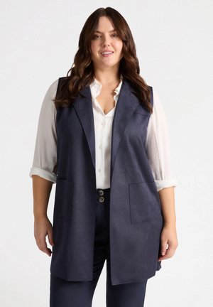 Navy mouwloze vest van faux suede met een gekartelde kraag, twee voorzakken en een open voorkant. Draagt over een witte blouse met knopen.