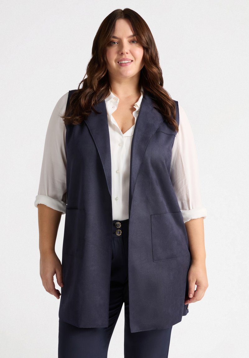 Marineblau ärmellose Weste aus Faux Wildleder mit Reverskragen, zwei Fronttaschen und offenem Vorderteil. Getragen über einem weißen Hemd mit Knöpfen.